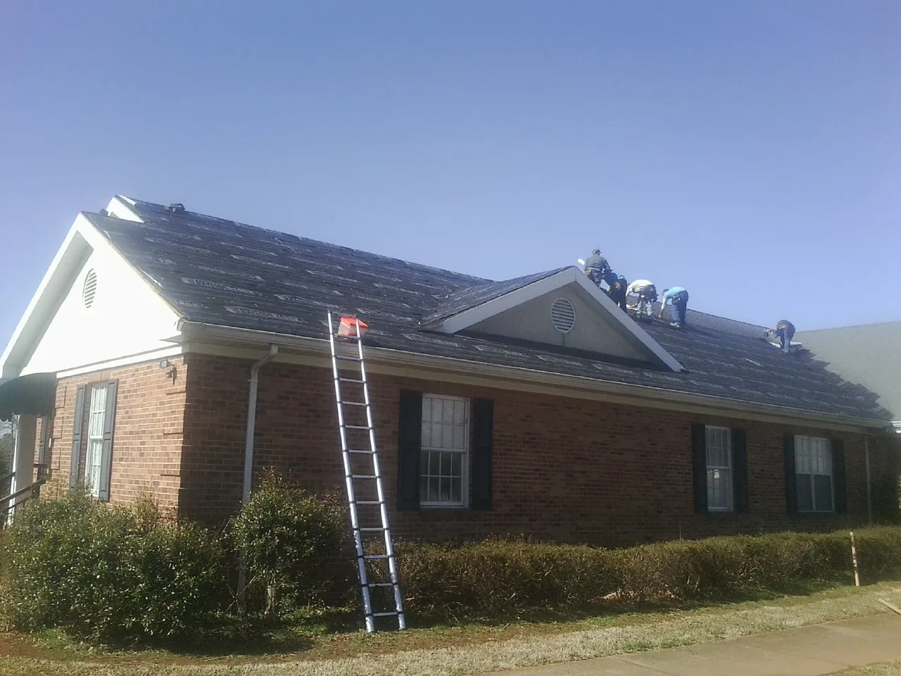 Crew tearing shingles off a brick ranch roof with a ladder set — Emergency Roof Repair work by our crew in Michie