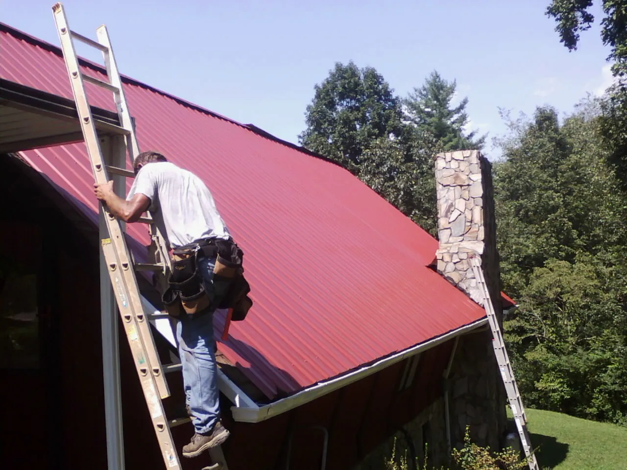 Roofer on a ladder beside a red metal roof and stone chimney — Soffit & Fascia Repair by Roofing Brothers in Michie